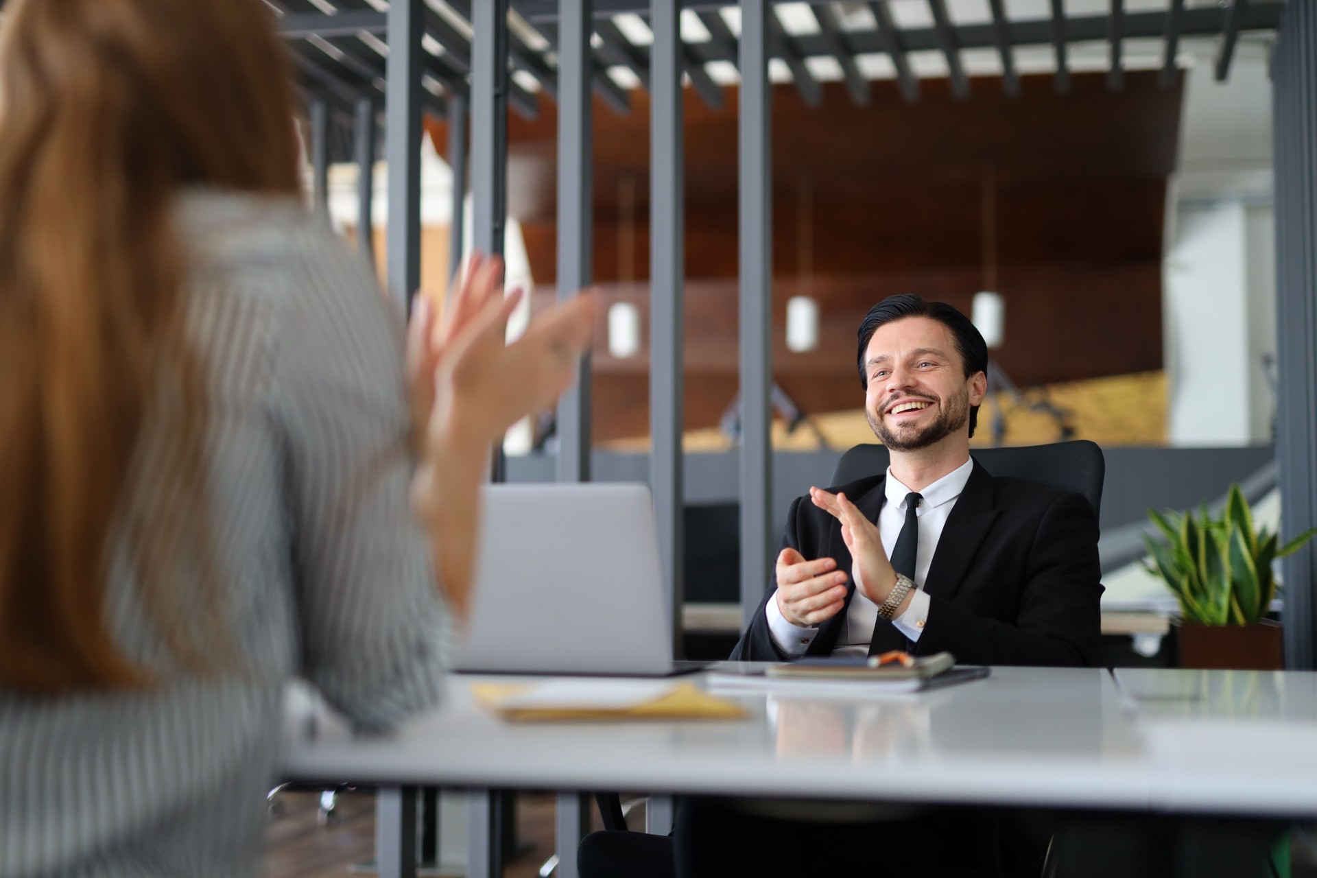 A man in a suit is sitting at a desk with a woman standing in front of him. The man is smiling and clapping his hands, while the woman is looking at him. The scene suggests a positive